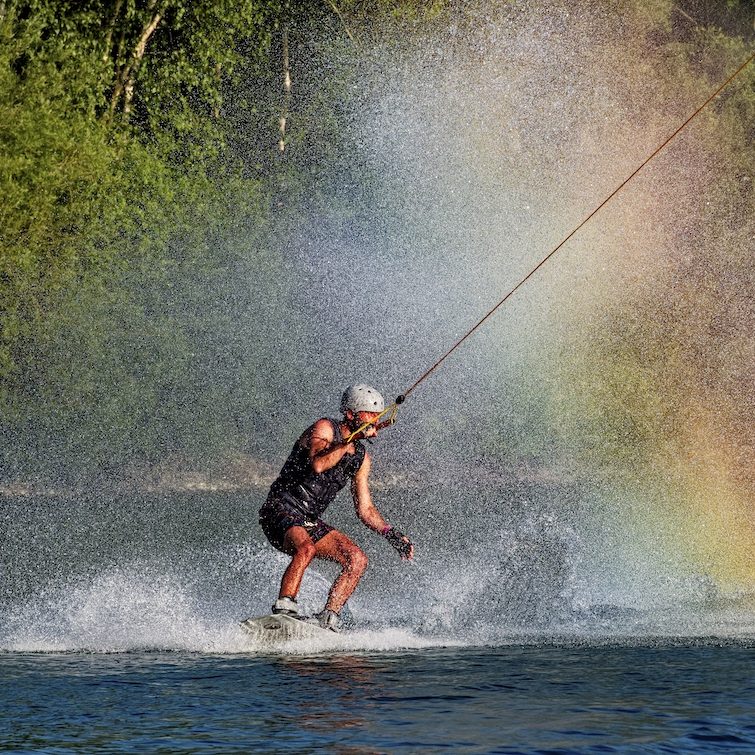 Wakeboarder springt über das Wasser vor einer sonnigen Berglandschaft.